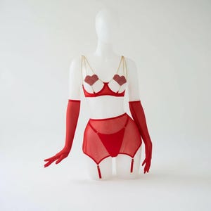 May include: Red lingerie set on a mannequin. The set includes a bra with heart-shaped cups, a garter belt, and a thong. The bra has gold chain straps. Long red gloves are also included.
