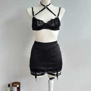 May include: Black lingerie set on a white mannequin. The set includes a bra with lace detailing, a halter neck, and a high-waisted skirt with garter straps. The bra has underwire support. The set is made of a shiny, satin-like material.