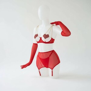 May include: Red lingerie set on a mannequin. The set includes a bra with heart-shaped embellishments, a garter belt, and matching gloves. The bra features gold chain straps. The lingerie is displayed against a white background.