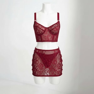 May include: A two-piece lingerie set in a deep burgundy hue. The set includes a bustier-style top with adjustable straps and a matching mini skirt. Both pieces are crafted from delicate lace, showcasing intricate floral patterns. The lingerie is displayed on a white mannequin.