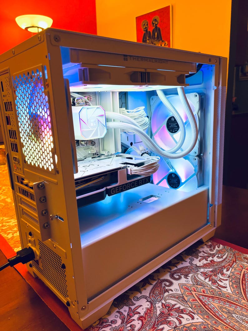 May include: A white computer case with internal components illuminated by blue and purple LED lights. The case features a graphics card labeled "GEFORCE RTX" and a CPU cooler. The text "THERMALRIGHT" is visible on the top of the case.
