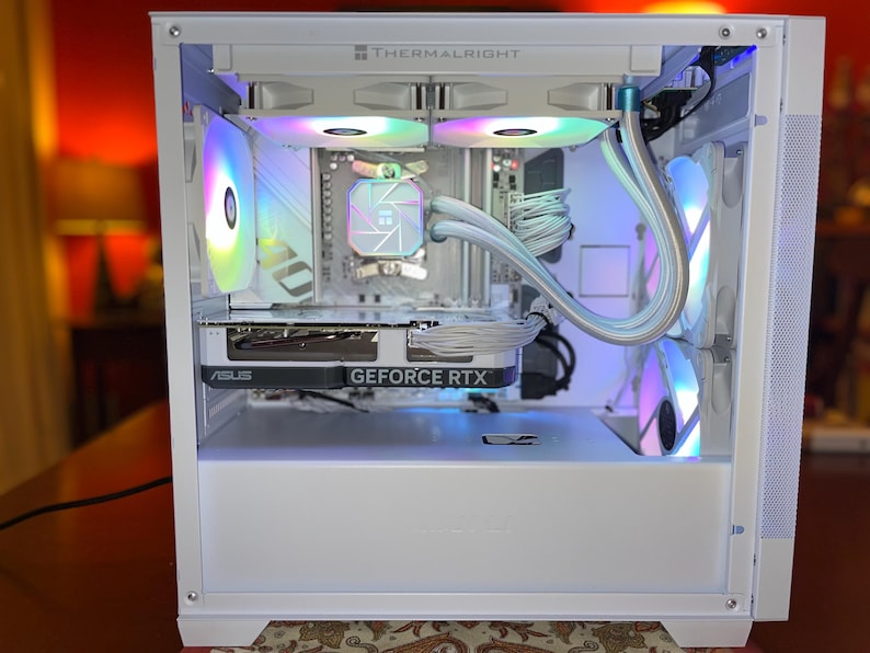 May include: White computer case with internal components, including a graphics card labeled "ASUS GEFORCE RTX." The case features multiple fans with colorful LED lighting. The top of the case is labeled "THERMALRIGHT."