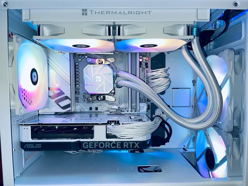 May include: A white computer case with internal components, including a graphics card labeled "GEFORCE RTX" and a CPU cooler. The case features multiple fans with colorful LED lighting. The brand "THERMALRIGHT" is visible at the top.