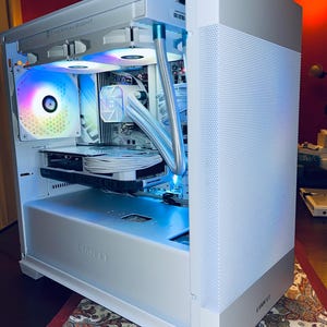 May include: A white computer case with a perforated side panel and internal components illuminated by colorful LED lights. The case features the "THERMALRIGHT" and "LIAN LI" brand names. The internal components include a graphics card and liquid cooling system.
