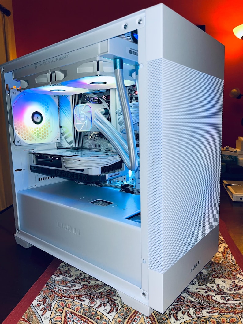 May include: A white computer case with a perforated side panel and internal components illuminated by colorful LED lights. The case features the "THERMALRIGHT" and "LIAN LI" brand names. The internal components include a graphics card and liquid cooling system.