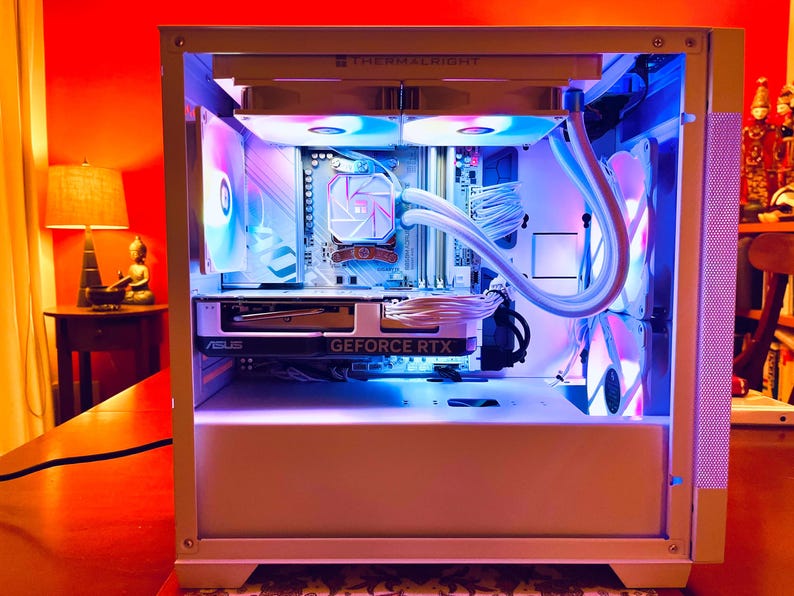 May include: A white computer case with internal components illuminated by blue and purple LED lights. The case features a graphics card with "GEFORCE RTX" printed on it. The components are neatly arranged, showcasing a modern PC build.
