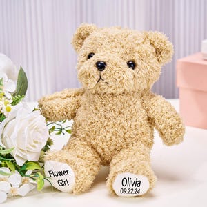 May include: A tan teddy bear with black eyes and a black nose. The bear has the words "Flower Girl" and "Olivia 09.22.24" printed on its feet. White flowers and a pink box are in the background.