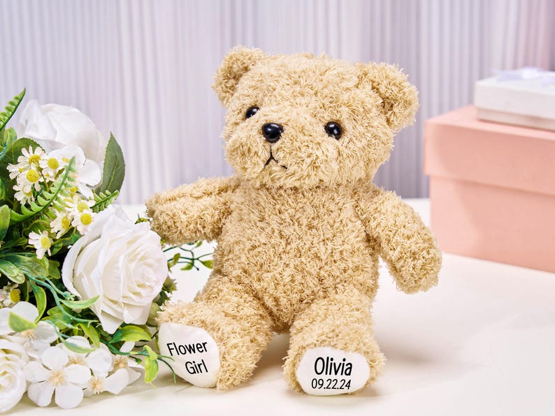 May include: A tan teddy bear with black eyes and a black nose. The bear has the words "Flower Girl" and "Olivia 09.22.24" printed on its feet. White flowers and a pink box are in the background.
