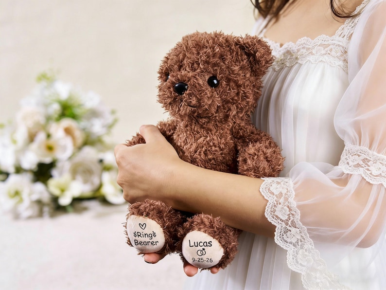 May include: A brown teddy bear with black eyes and a textured fur appearance. The bear has the words "Ring Bearer" and "Lucas 9-25-26" printed on its paws. The bear is held by a person wearing a white dress.