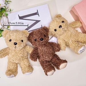 May include: Three plush teddy bears in shades of tan and brown, each approximately 9.84 inches tall. One bear is a darker brown. The bears are lying on a surface with a white card and a pink box in the background.