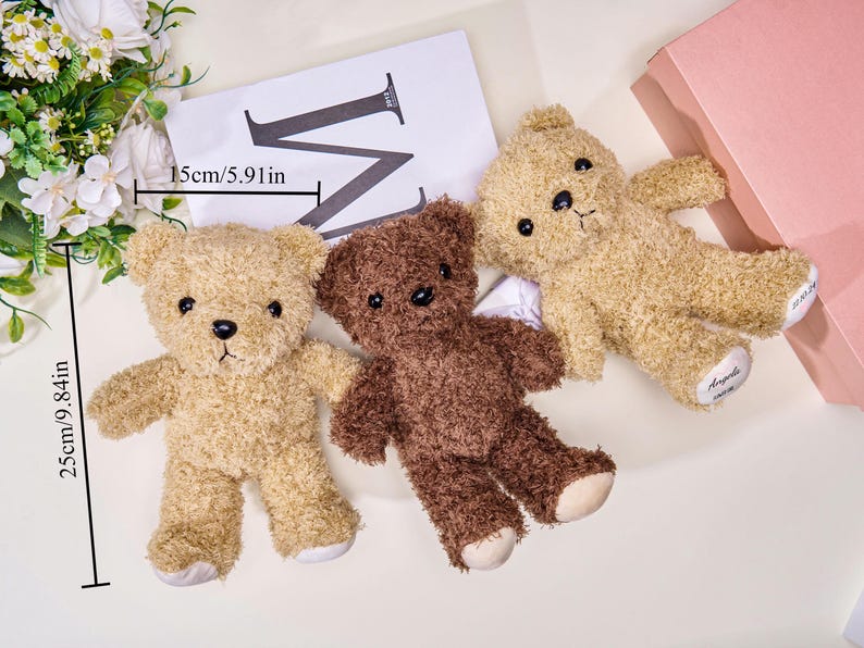 May include: Three plush teddy bears in shades of tan and brown, each approximately 9.84 inches tall. One bear is a darker brown. The bears are lying on a surface with a white card and a pink box in the background.