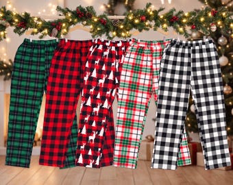 Puede incluir: Cinco pantalones de estar por casa colgados en perchas de madera. Los pantalones presentan varios estampados, incluyendo cuadros verdes y negros, cuadros rojos y negros, un fondo negro con renos y árboles blancos, cuadros rojos, verdes y blancos, y cuadros negros y blancos.