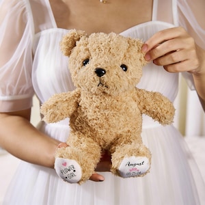 May include: A plush, light brown teddy bear with a curly texture. The bear has black eyes and a black nose. The left foot has the text "FLOWER GIRL" and a heart and star. The right foot has the name "August" and the date "08-25-24".