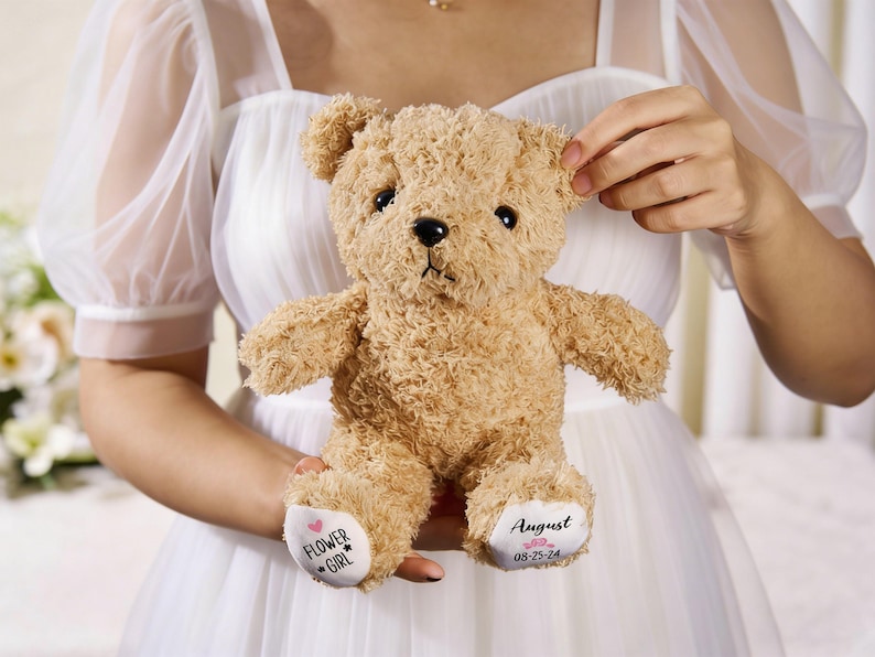 May include: A plush, light brown teddy bear with a curly texture. The bear has black eyes and a black nose. The left foot has the text "FLOWER GIRL" and a heart and star. The right foot has the name "August" and the date "08-25-24".
