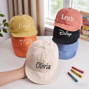 Op de afbeelding: Een stapel baseballpetten in verschillende kleuren, waaronder geel, oranje, perzik, zwart en blauw. Elke pet heeft een geborduurde naam: Bruce, Tyler, Lewis, Daisy en Olivia. Er liggen kleurpotloden op tafel.