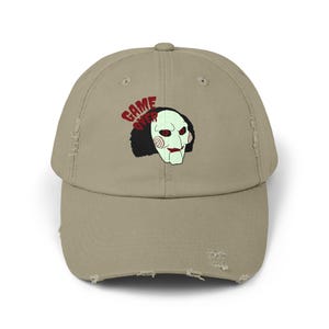 Könnte beinhalten: Eine khaki-farbene, abgenutzte Baseballkappe mit einer Grafik einer weißen Maske mit roten Akzenten und schwarzem Haar. Die Worte "GAME OVER" sind rot über der Maske aufgedruckt. Die Kappe hat einen gebogenen Schirm und ein abgenutztes Aussehen.