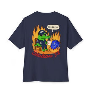 以下が含まれることがあります： FDNYの帽子をかぶった漫画のアリゲーター消防士が、炎に囲まれた椅子に座っているネイビーブルーのTシャツ。「THIS IS FINE」というテキストが吹き出しの中にあります。「Station 32」という言葉は赤色です。