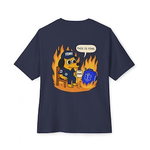 Op de afbeelding: Marineblauw T-shirt met een cartoonhond in een FDNY EMS-uniform, zittend in vlammen, met een mok. Er is ook een EHBO-kit aanwezig. De tekst "THIS IS FINE" verschijnt in een tekstballon.