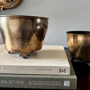 May include: Two antique brass-toned metal bowls with ornate feet rest on a stack of books. The larger bowl sits atop a book with the title "Provençal", while the smaller bowl is beside it.