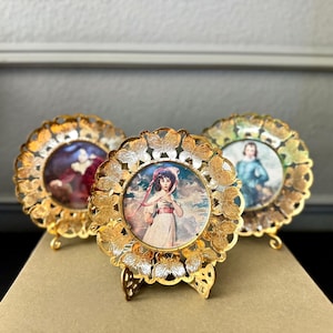 May include: Three antique gold-colored picture frames with ornate butterfly-shaped designs. Each frame holds a small, vintage portrait. The frames are displayed on a neutral surface, showcasing their intricate details and aged appearance.