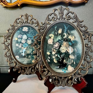 May include: Two antique oval picture frames with floral still life prints. The frames have ornate, bronze-colored filigree detailing and are displayed on dark wooden stands. The prints feature bouquets of flowers in vases, with soft blues, pinks, and greens.
