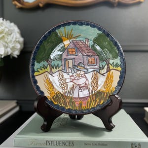 May include: A decorative ceramic plate featuring a hand-painted rural scene with a house, trees, and a figure in a field. The plate is displayed on a wooden stand, showcasing its colorful design and intricate details. The plate has a dark blue border.