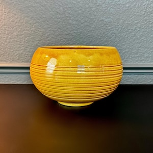 May include: A round, mustard yellow ceramic bowl featuring a glossy finish and a series of horizontal, recessed grooves encircling the lower half of the exterior. The bowl has a wide, open top and a smooth, rounded silhouette with a small base.
