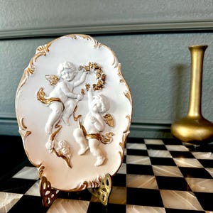 May include: An oval white ceramic plaque featuring two raised cherub figures in relief. The cherubs are accented with gold paint on their wings, hair, and a floral wreath they hold. The plaque has a decorative gold-painted scalloped edge.