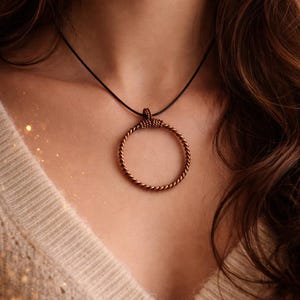 May include: A copper-colored, circular pendant necklace. The pendant is a twisted wire design, suspended from a black cord. The necklace is worn against a beige sweater. The image is a close-up, focusing on the necklace and upper chest.