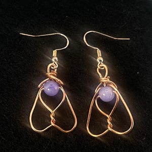 May include: A pair of handmade earrings featuring a light purple bead suspended within a copper-colored wire frame. The wire is shaped into a stylized wing or heart design, with a simple hook for the ear.