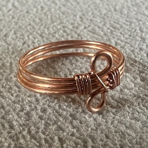 May include: A handcrafted copper wire ring with a unique design. The ring features multiple bands of copper wire, with a decorative looped accent. The ring is displayed on a textured, light-colored surface.