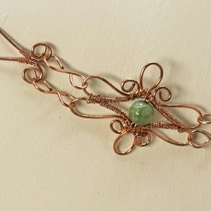 May include: A copper wire bracelet featuring intricate hand-formed loops and swirls. The design incorporates a single round, pale green gemstone bead secured in the center by coiled wire wrapping, with a simple hook-and-loop closure mechanism.