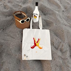 May include: A beach scene with a brown sun visor with cream sunglasses, a bottle of Malibu rum, and a canvas tote bag. The bag has a graphic of two wine glasses with red and orange liquid. The items are on sand.