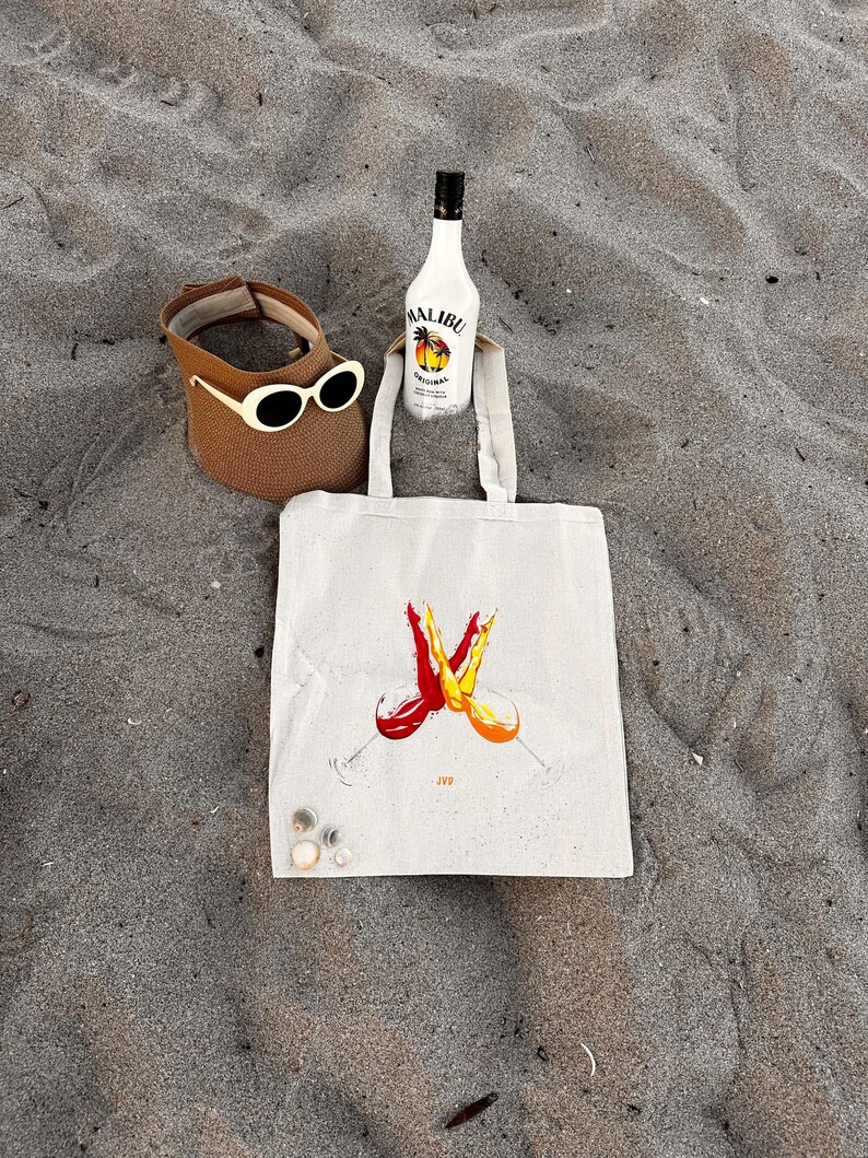 May include: A beach scene with a brown sun visor with cream sunglasses, a bottle of Malibu rum, and a canvas tote bag. The bag has a graphic of two wine glasses with red and orange liquid. The items are on sand.