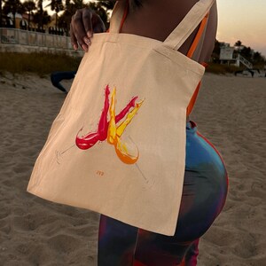 May include: A beige tote bag featuring a watercolour design of two wine glasses, one filled with red wine and the other with orange wine. The bag has long, light-coloured straps and the initials "JVD" are printed on the bag.