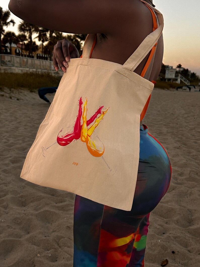 May include: A beige tote bag featuring a watercolour design of two wine glasses, one filled with red wine and the other with orange wine. The bag has long, light-coloured straps and the initials "JVD" are printed on the bag.