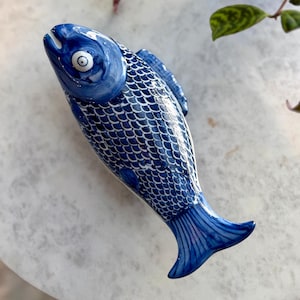 May include: A ceramic fish figurine in shades of blue, featuring a detailed scale pattern and a white eye. The fish has a curved body and a flared tail. The figurine is placed on a white marble surface, with a potted plant in the background.