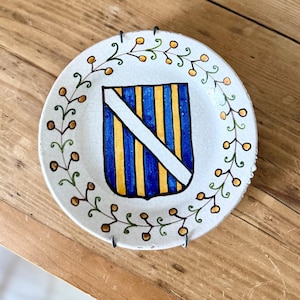 May include: A small, round, white ceramic plate with a hand-painted design. The plate features a blue and yellow striped shield with a white diagonal stripe. A green and brown floral border encircles the shield. The plate is displayed on a wooden surface.
