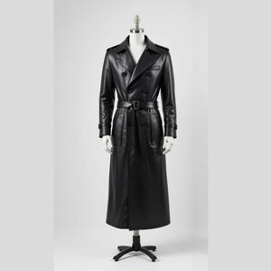 Puede incluir: Un abrigo largo de cuero negro, tipo gabardina, exhibido en un maniquí. El abrigo tiene un diseño de doble botonadura, cinturón en la cintura y cuello clásico. El abrigo es largo y tiene mangas largas.