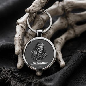 May include: A round, silver keychain with a black pendant featuring a skull wearing a beanie and the text "I AM IMMORTAL". The skull is giving a hand gesture. The words "BIGS & GRIM" are above the skull.