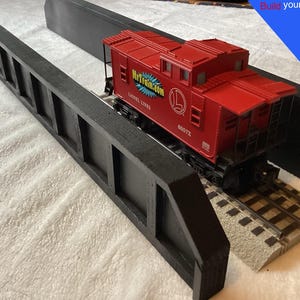 May include: A red Lionel Lines caboose on a model train track, positioned between two black bridge supports. The caboose has the text "MrTrain.com" and the number "6057X". A blue banner in the background reads "Build your own bridge".