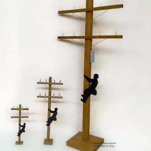 May include: Wooden lineman decorations in three sizes. Each features a brown wooden utility pole with cross arms and a black silhouette of a lineman climbing. The largest pole has a gray box attached. "Lineman Decorations at MrTrain on Etsy" is printed on the bottom right.