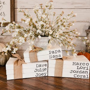 May include: A stack of white books tied with burlap ribbon, featuring the names Papa, Nana, Dave, Julie, Joey, Marco, Lori, Jordan, and Coral printed in black on the spines. The books are arranged in three separate stacks.
