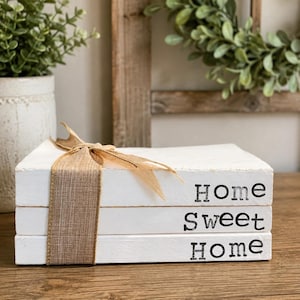 May include: A stack of three rectangular white wooden blocks resembling books, tied together with a tan burlap ribbon. The words Home, Sweet, and Home are printed in black ink on the side of each block, reading from top to bottom.