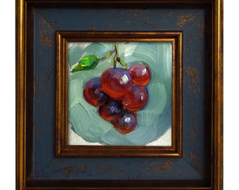 May include: Oil painting of a cluster of red grapes with a green leaf, set against a light blue background. The artwork is framed in a square, dark blue frame with a gold inner edge, creating a classic look.