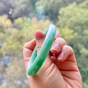 May include: A smooth, polished bangle bracelet crafted from translucent green jade. The circular accessory features natural color variations with deep emerald green swirls blended into a lighter, pale mint green base, creating a glossy finish.