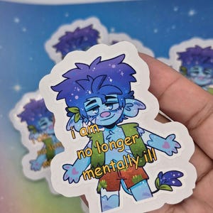 Puede incluir: Una pegatina con un personaje de dibujos animados azul con el texto "I am no longer mentally ill". El personaje tiene piel azul, pelo morado y viste ropa verde y naranja. Hay pegatinas adicionales en el fondo.