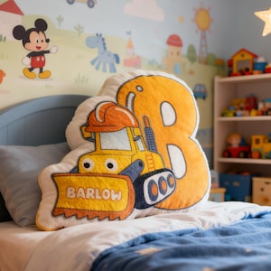 May include: A yellow and orange bulldozer pillow with the name "Barlow" on the blade, and a large number "8" pillow. The pillows are on a bed with a blue comforter. The room has a cartoon mural and a star-shaped light.