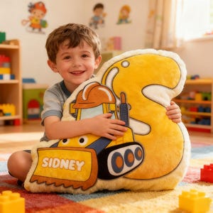 May include: A plush yellow pillow in the shape of the number 3, featuring a cartoon bulldozer design with the name "SIDNEY" on the front. The pillow is held by a smiling child in a playroom setting.