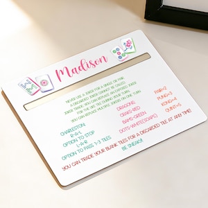 May include: A white Mahjong tile holder with the name "Madison" in pink script. The holder has a slot and features colorful illustrations of Mahjong tiles and game instructions in various colours.
